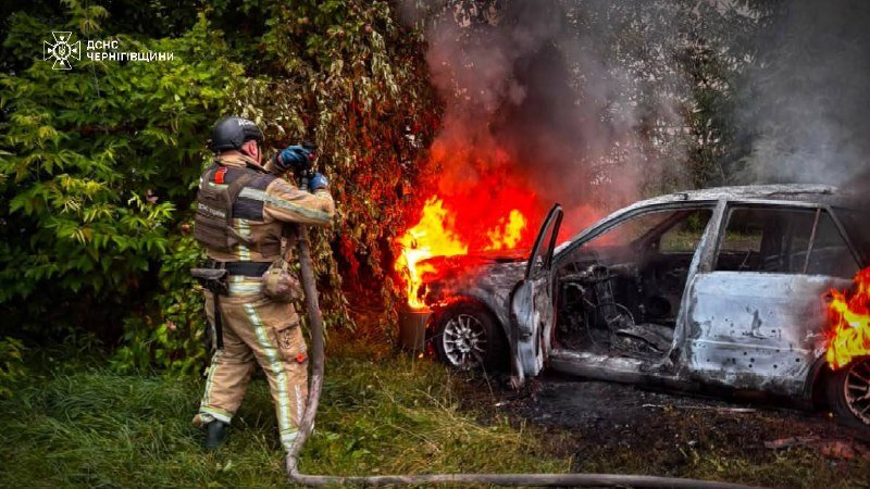 A firefighter in protective gear sprays water from a hose onto a burning car with its driver's door open, surrounded by flames and smoke.