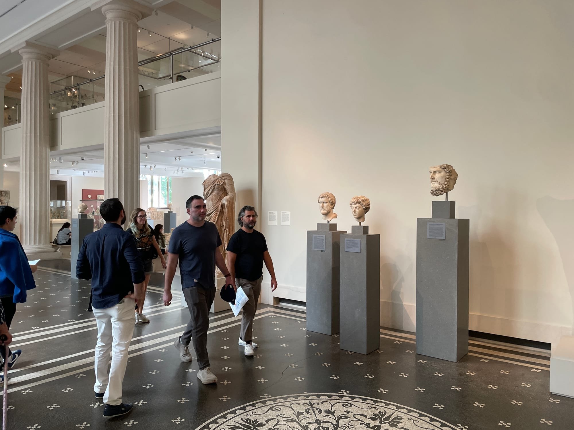 Visitors walk through a museum gallery featuring classical marble sculpture heads displayed on pedestals beneath tall columns.