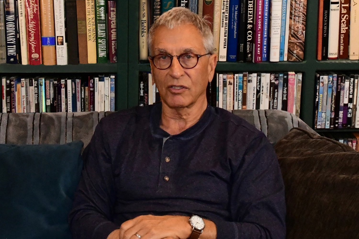 A gray-haired man wearing glasses and a dark henley shirt sits on a couch in front of a bookshelf filled with books and DVDs.