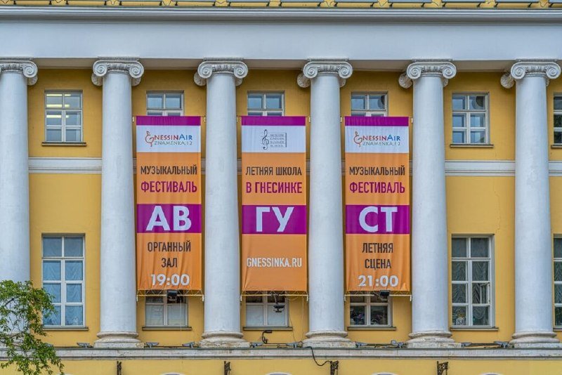 Three vertical banners advertising "GnessinAir" festival events hang between the white columns of a yellow classical building facade.