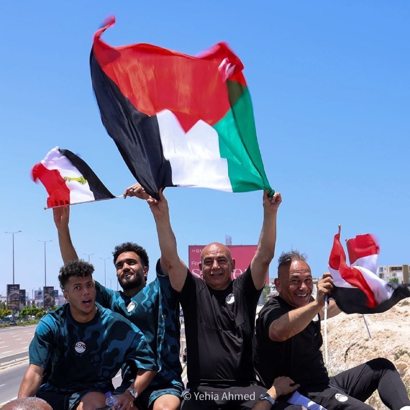 Four men in dark athletic shirts smile and wave flags outdoors on a sunny day near a roadside with billboards in the background.