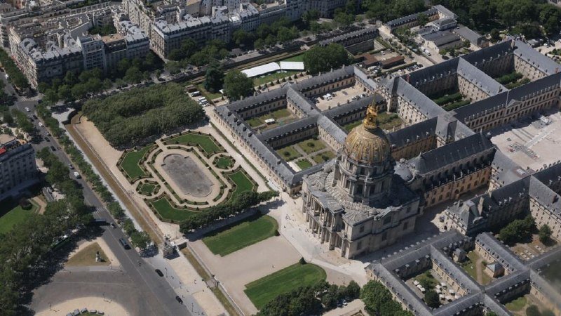 Aerial view of a large stone building with a gilded dome, surrounded by formal gardens, courtyards, and adjacent urban blocks with trees lining the streets.