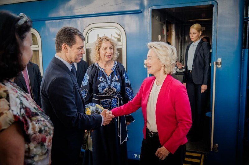 A woman in a red blazer shakes hands with a man in a dark suit beside a blue train, as others watch and one stands inside the train doorway.