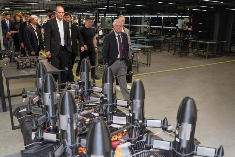 A group of people in business attire walk through a large industrial workshop past rows of black missile-like devices arranged on tables.