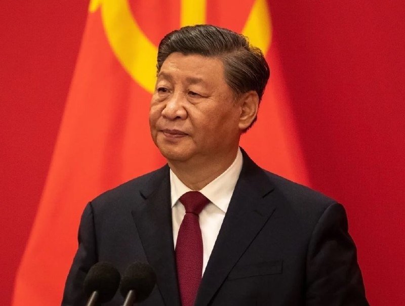 A man in a dark suit and red tie stands at a podium with microphones, positioned before red flags displaying a yellow hammer and sickle emblem.