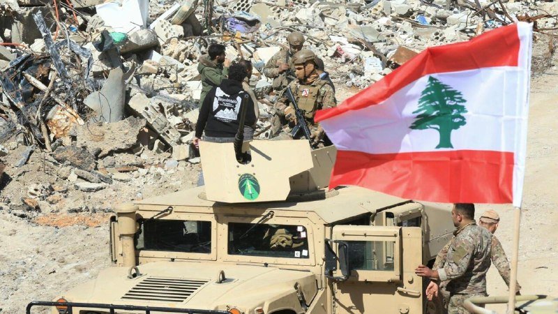Armed soldiers in camouflage uniforms ride atop a tan armored vehicle past concrete rubble, with a large red-and-white Lebanese flag displayed prominently on the vehicle.