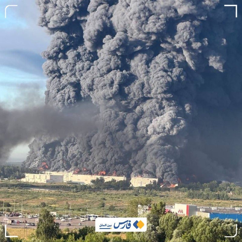 A massive column of thick black smoke rises from a large industrial building engulfed in visible flames, towering over a rural roadside landscape with an Arabic news watermark in the corner.