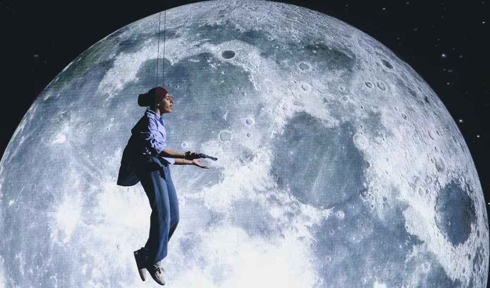 A performer suspended by wires holds a microphone against a large projected moon backdrop.