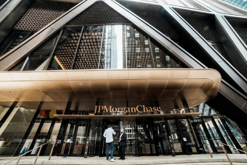 Two people stand talking beneath a curved metallic awning marked "JPMorganChase" at the glass entrance of a modern, angular high-rise building.