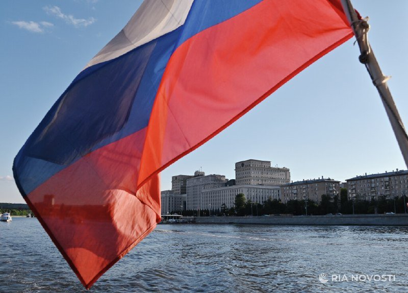 A Russian tricolor flag waves in the foreground against a river, with city buildings and a waterfront visible in the background.