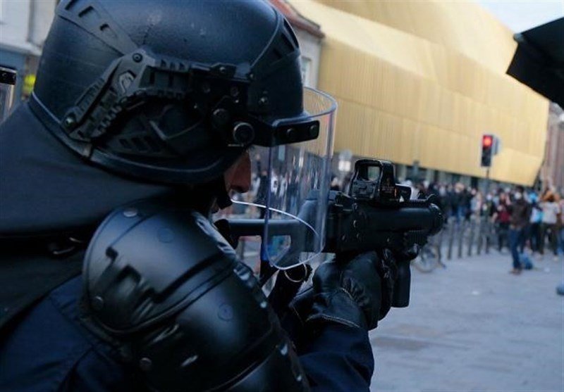 A helmeted officer aims a less-lethal launcher toward a crowd on a city street.