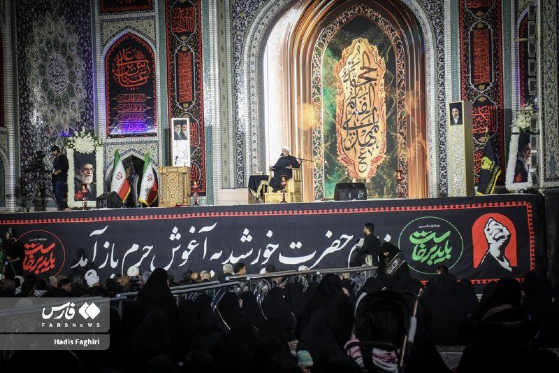 A speaker sits on stage at an ornate religious shrine addressing a seated crowd, flanked by framed portraits and Persian-language banners.