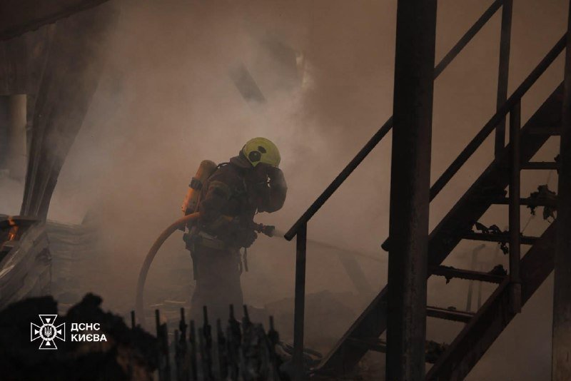 A firefighter in yellow helmet and gear walks through a smoke-filled, fire-damaged interior near a metal staircase, with "ДСНС КИЄВА" logo visible.