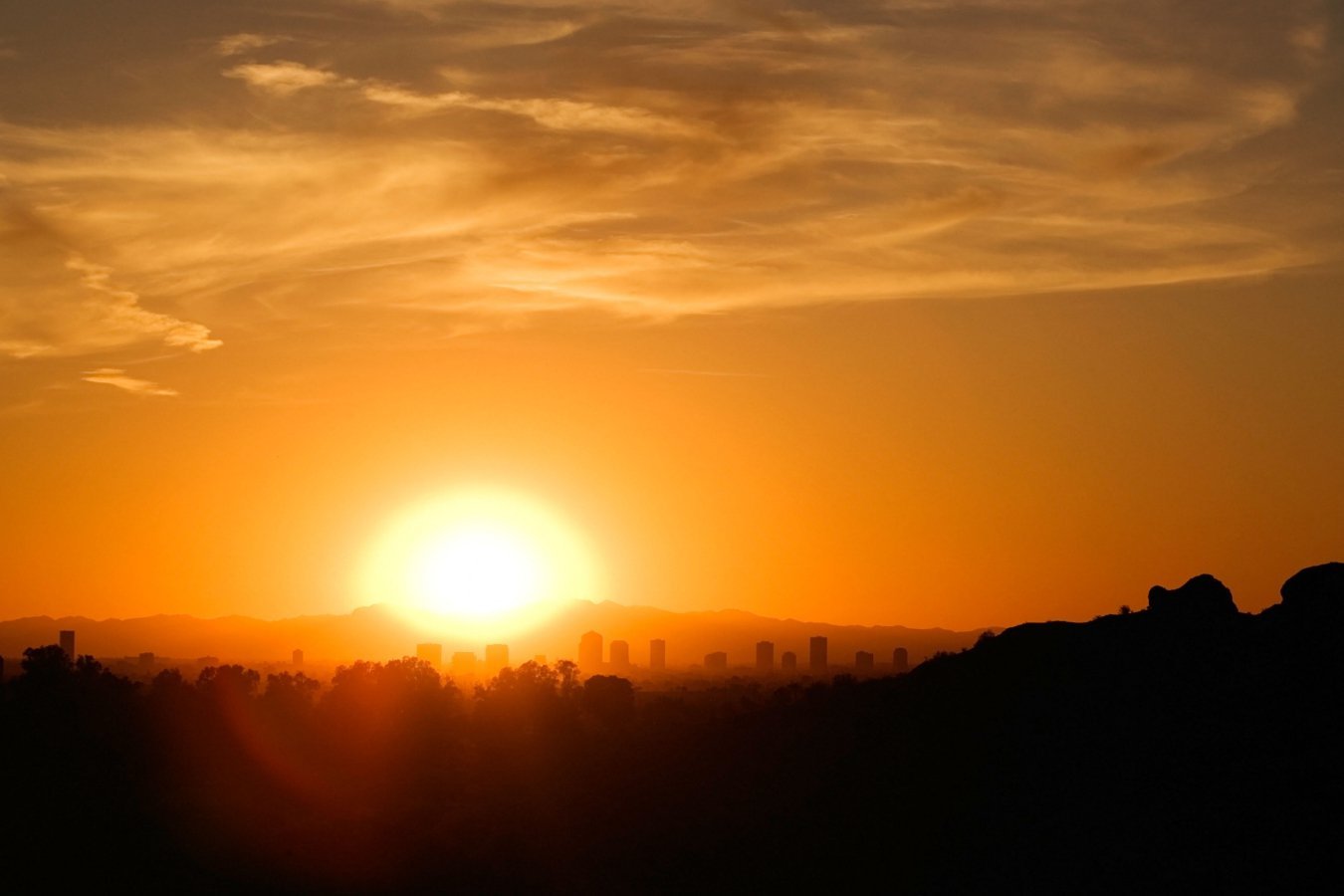 The sun sets behind a silhouetted city skyline, with orange and golden light illuminating scattered clouds above distant mountains and dark hills.