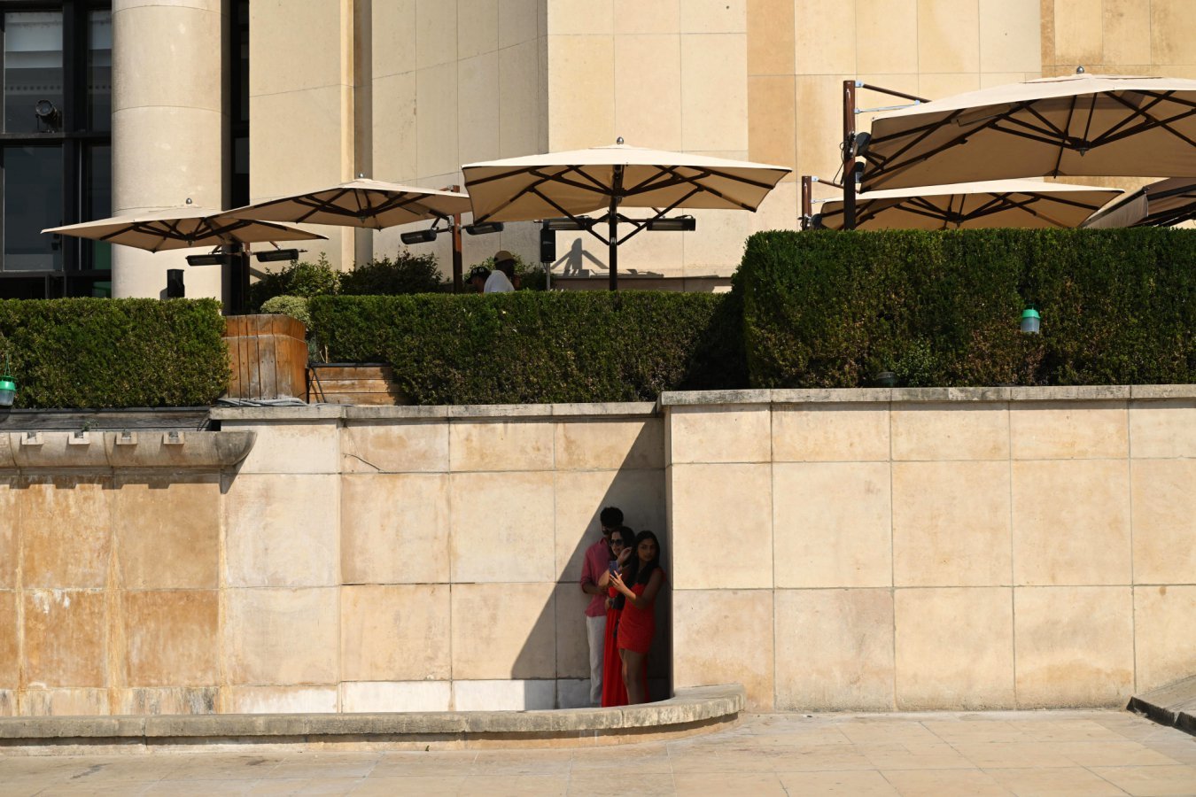 Three people stand together in a shaded alcove of a stone building, holding up a phone beneath large patio umbrellas and neatly trimmed hedges.