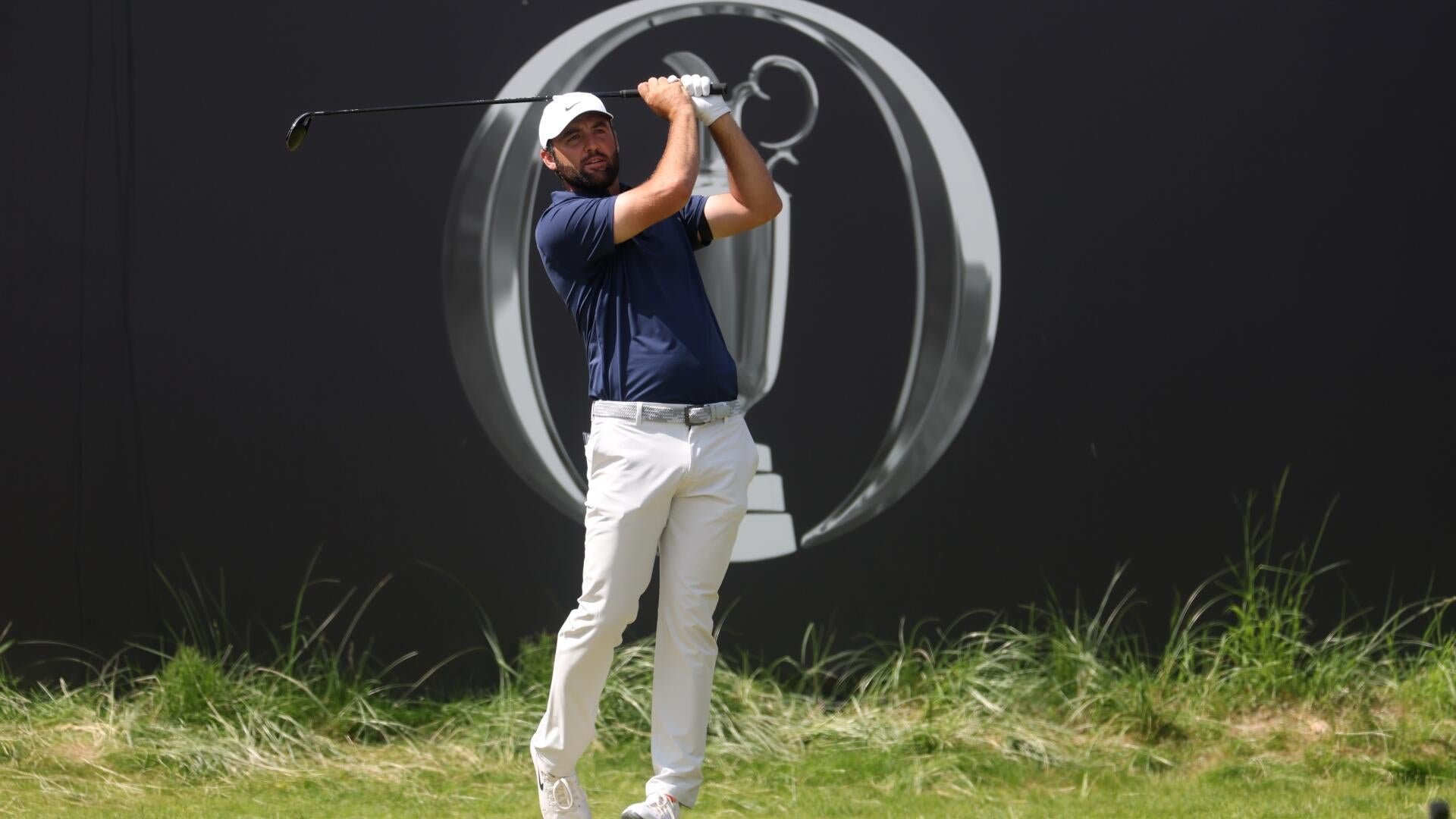 A bearded golfer in a navy shirt, white pants, and white cap completes a swing with a driver, positioned on grass in front of a black backdrop displaying a trophy logo.