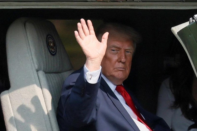 A man in a dark suit and red tie waves from the interior of a vehicle bearing a presidential seal.