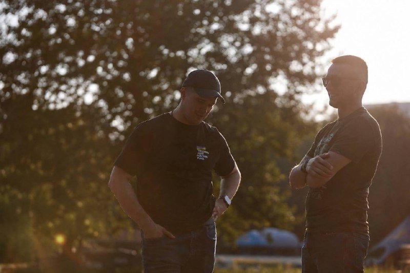 Two men in dark t-shirts stand outdoors against a sunlit, tree-filled backdrop, one wearing a cap and looking down, the other in sunglasses with arms crossed.
