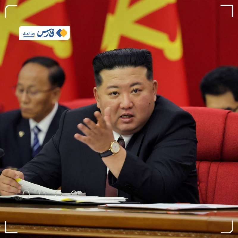 A man in a dark suit gestures with his hand while seated at a table with documents, speaking in front of a red and yellow emblem with two other men partially visible nearby.