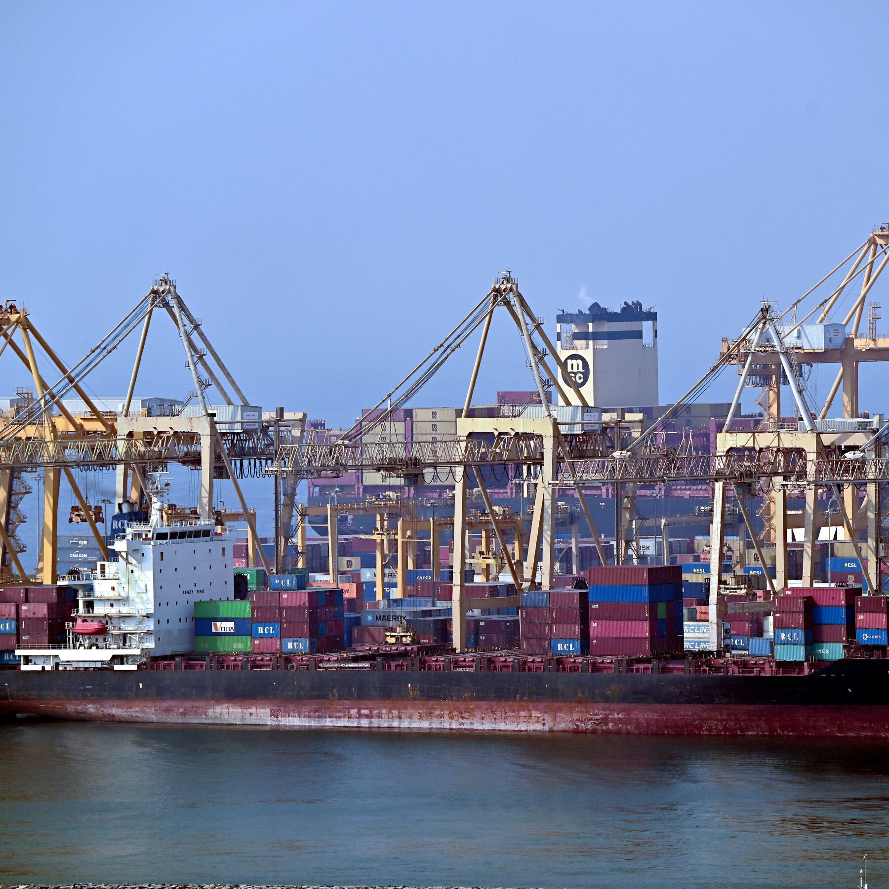A large container ship loaded with multicolored shipping containers is docked at a busy port, with multiple tall cranes towering over stacked containers in the background.