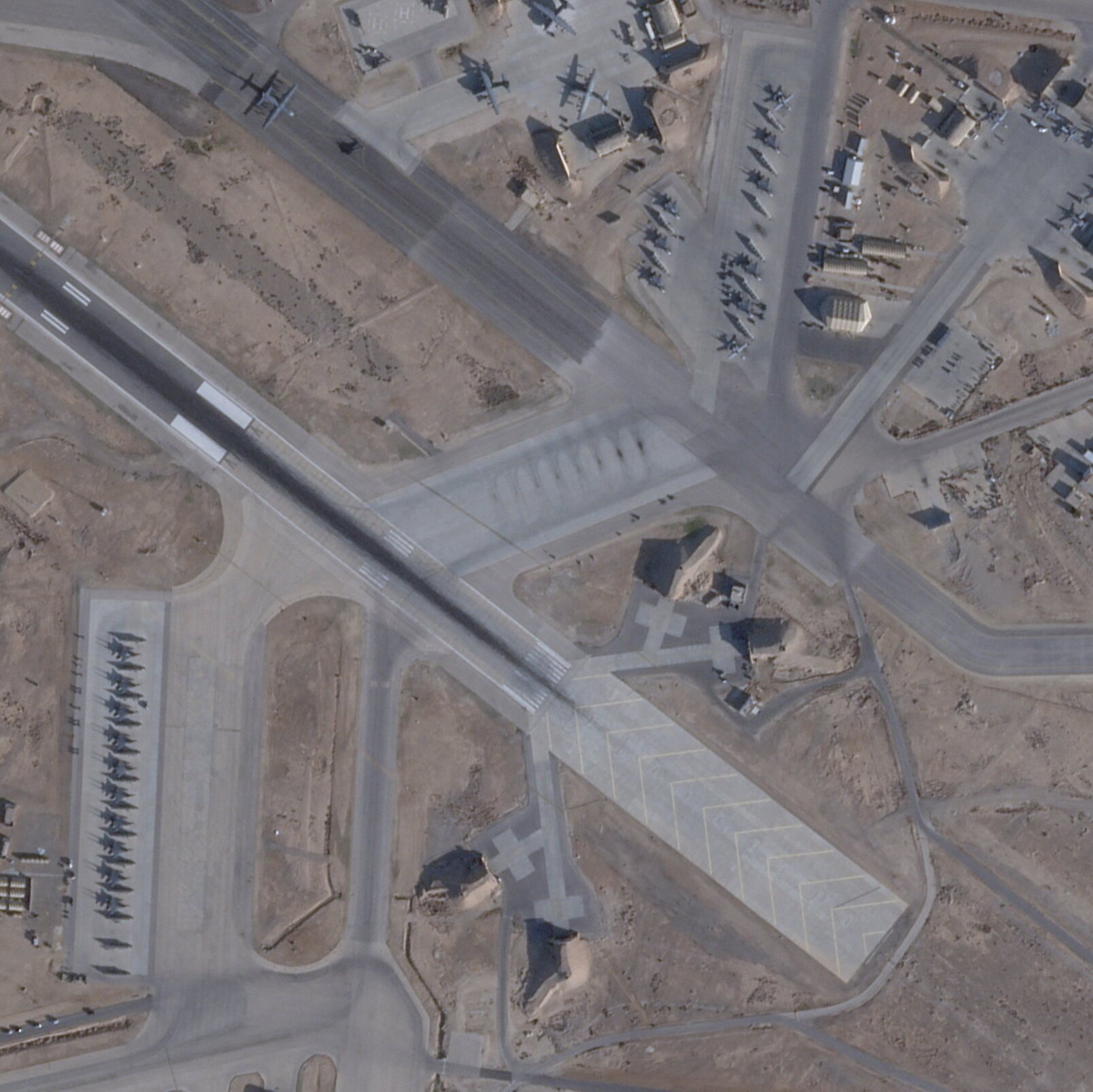 Satellite image showing a desert airfield with multiple military aircraft parked on aprons alongside a runway, taxiways, revetments, and hangars.