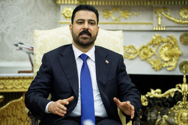 A bearded man in a dark suit and blue tie, wearing a flag lapel pin, sits in an ornate cream chair gesturing with both hands in a gilded, formal room.