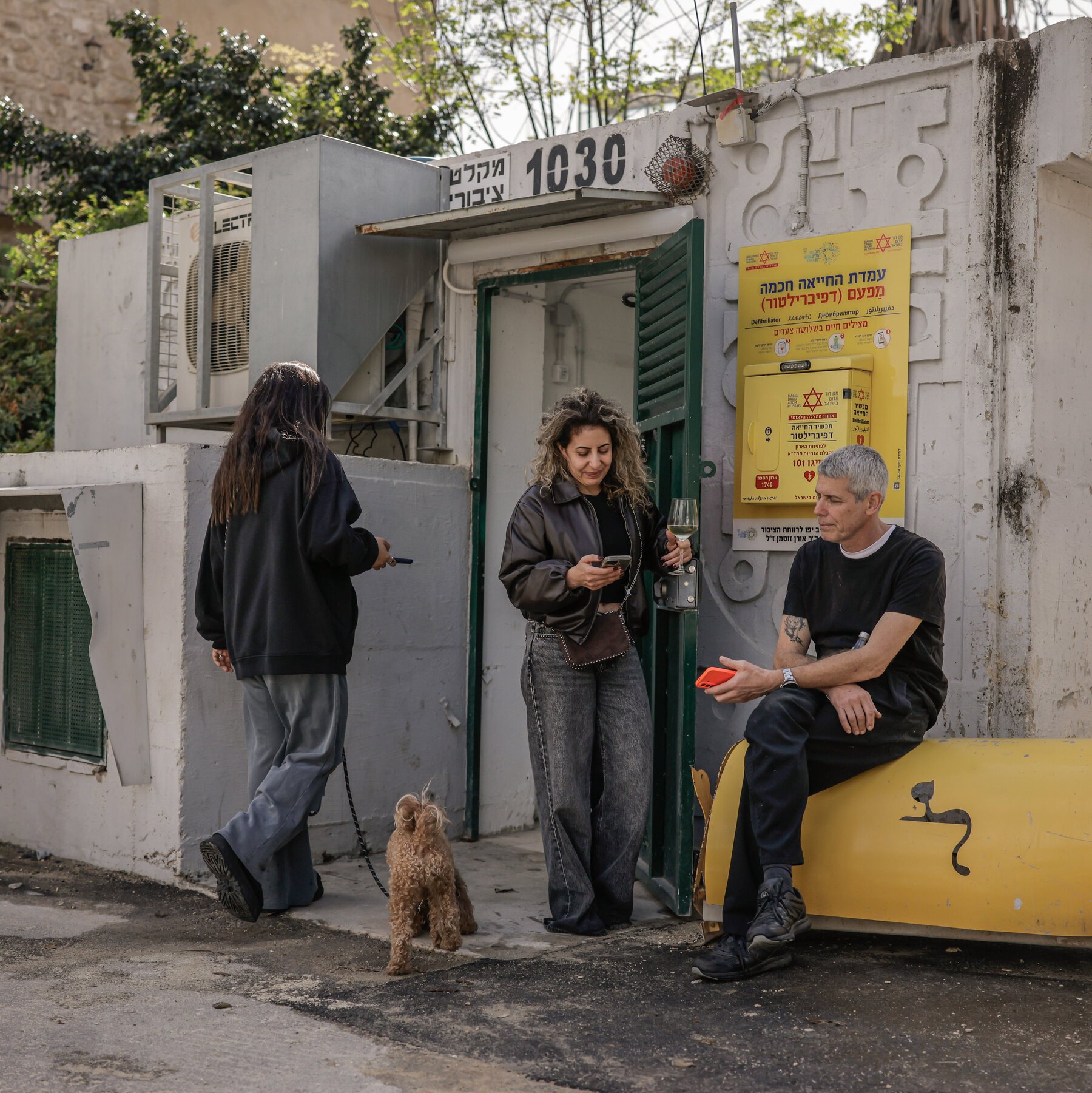 Three people gather near a yellow defibrillator station: a woman with a small dog on a leash, a woman holding a wine glass and phone, and a seated man with a red phone.
