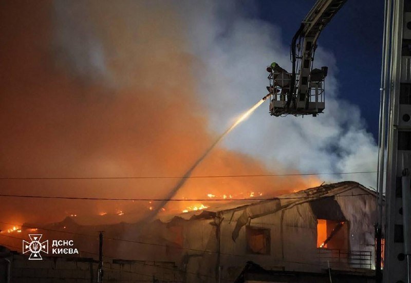 Firefighters in an aerial ladder bucket spray water onto a burning building at night, with flames and thick smoke visible; a logo reads "ДСНС КИЄВА."