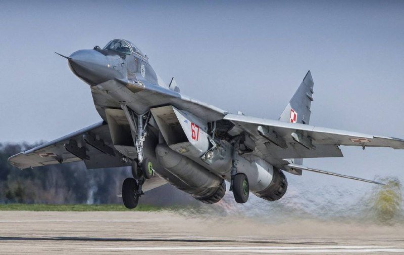 A gray military fighter jet with red and white checkered markings and the number "67" lifts off from a runway, landing gear extended.