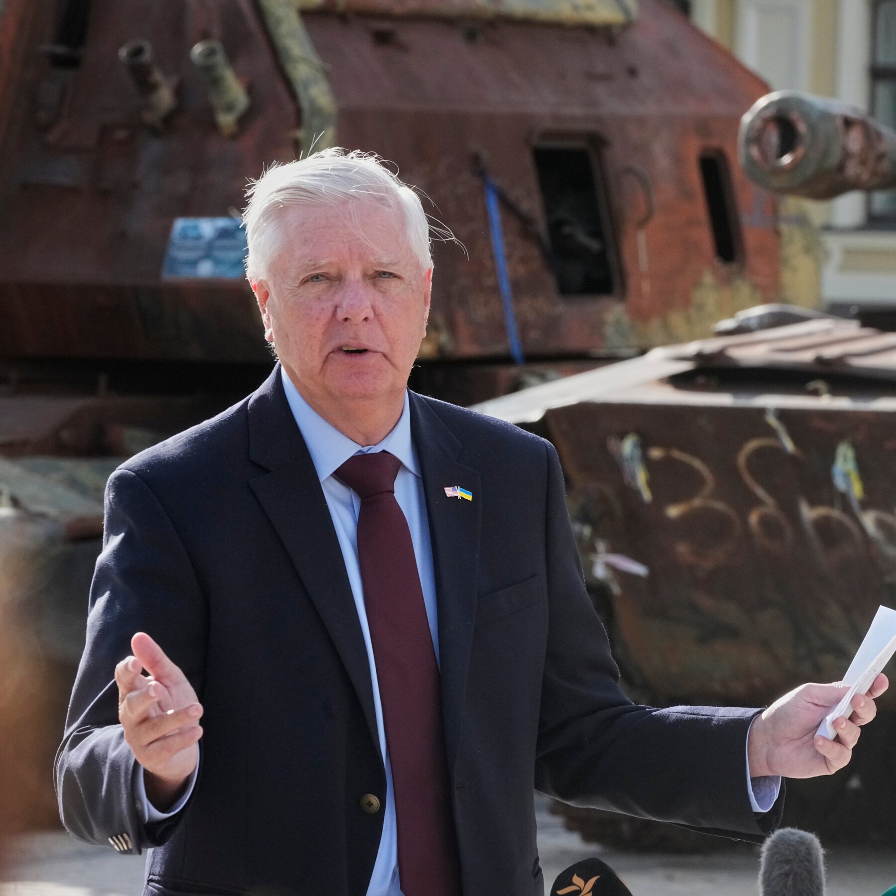 A white-haired man in a dark suit with US and Ukrainian flag lapel pins speaks while gesturing, holding a paper, with rusted military tanks visible behind him.