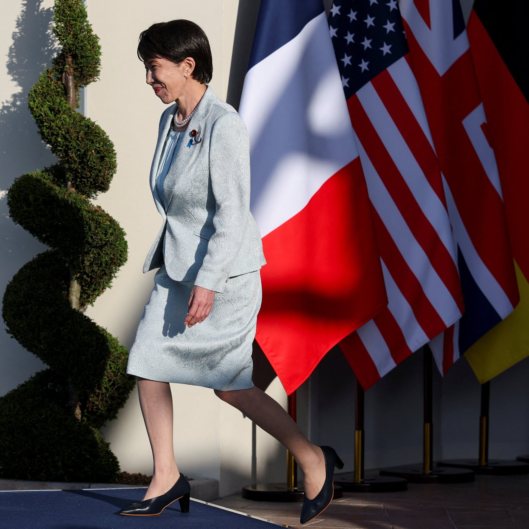 A woman in a light blue suit walks down steps past the American flag and other national flags displayed outdoors.