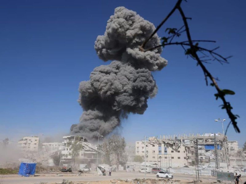 A large plume of gray smoke rises above partially destroyed buildings, with dust, scattered debris, vehicles, and a few people visible on the ground below.
