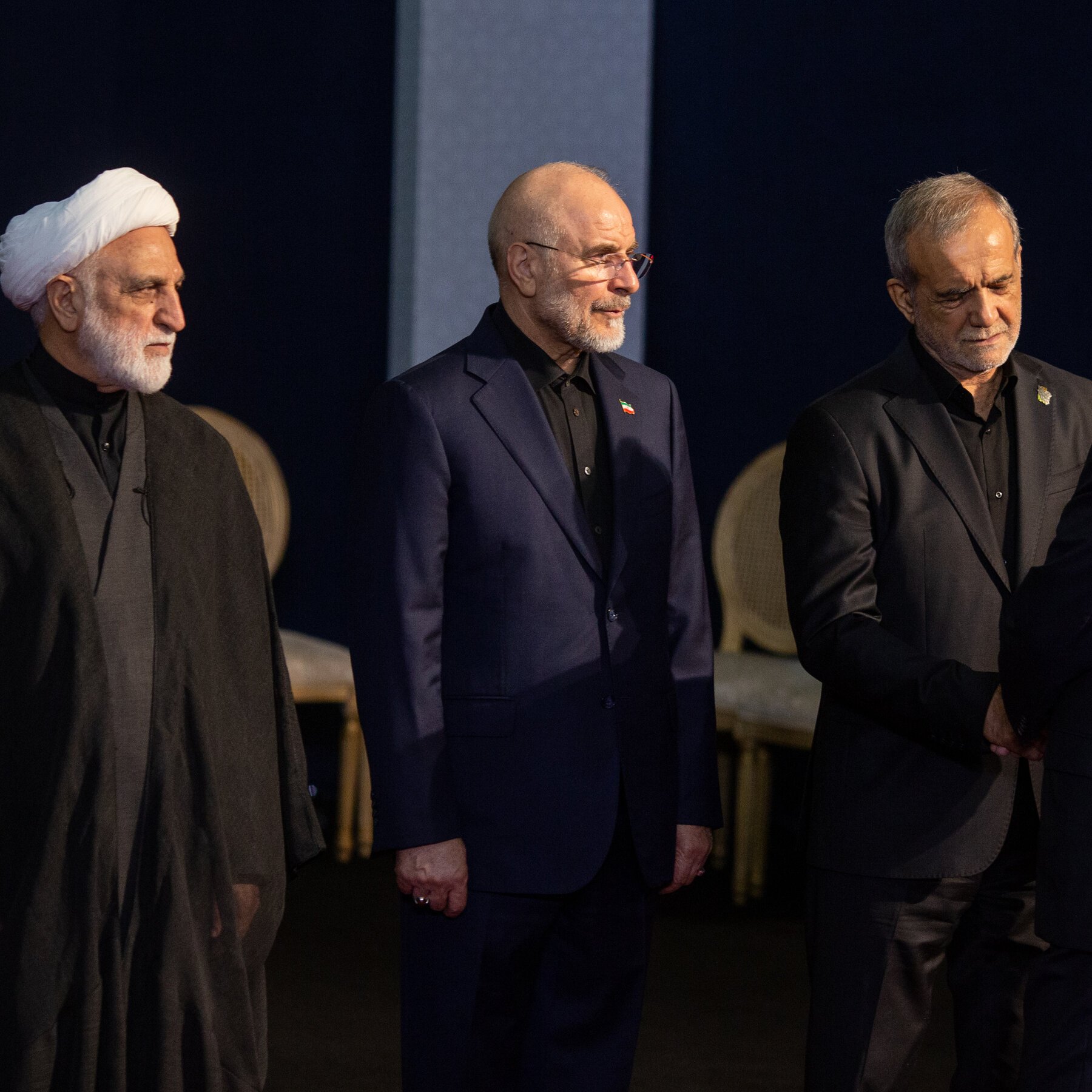 Three older men in dark suits and traditional clerical attire stand solemnly on a dimly lit stage with empty chairs behind them.