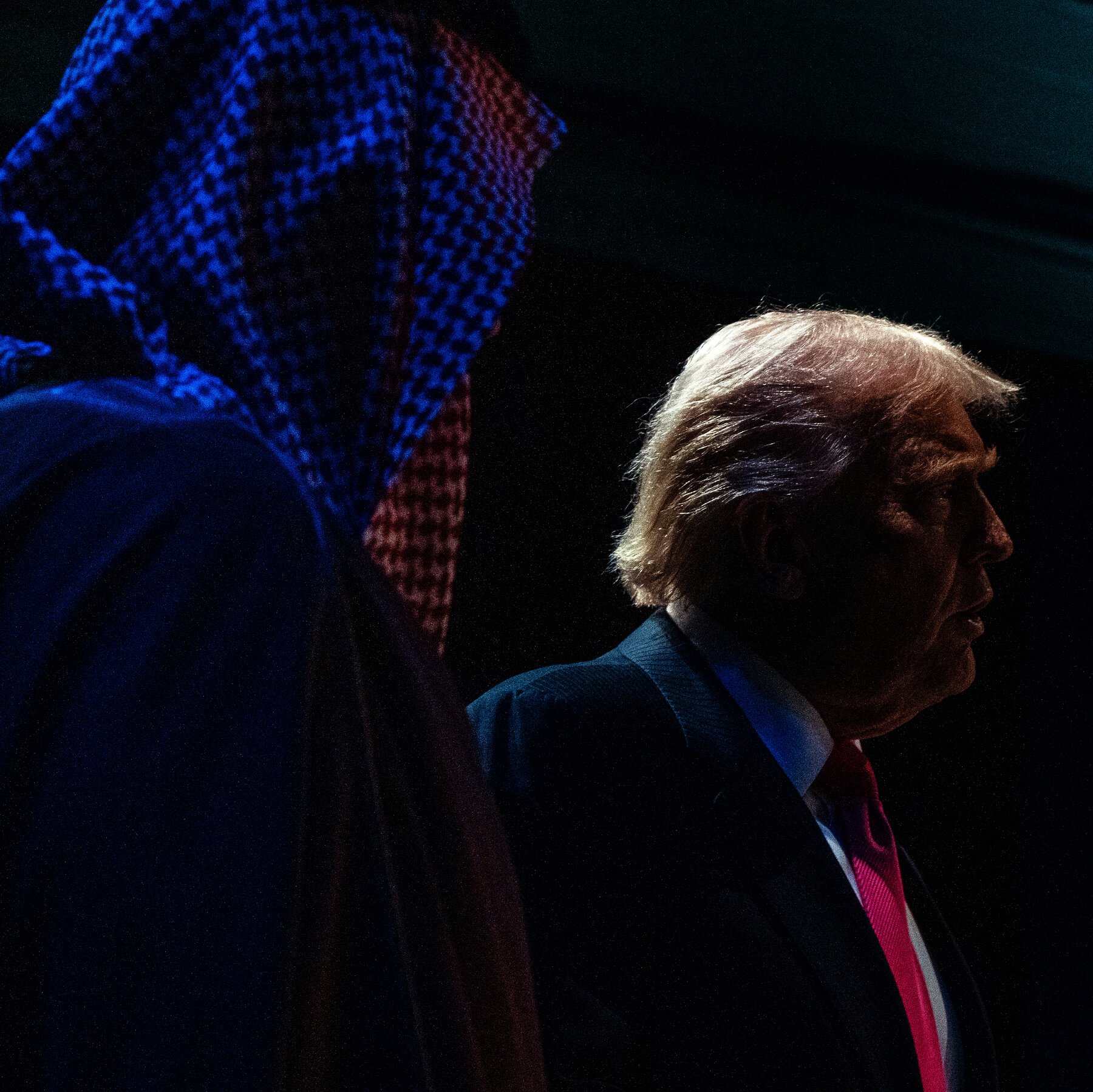 A man in a dark suit and red tie walks past a dimly lit American flag, his face shown in profile.