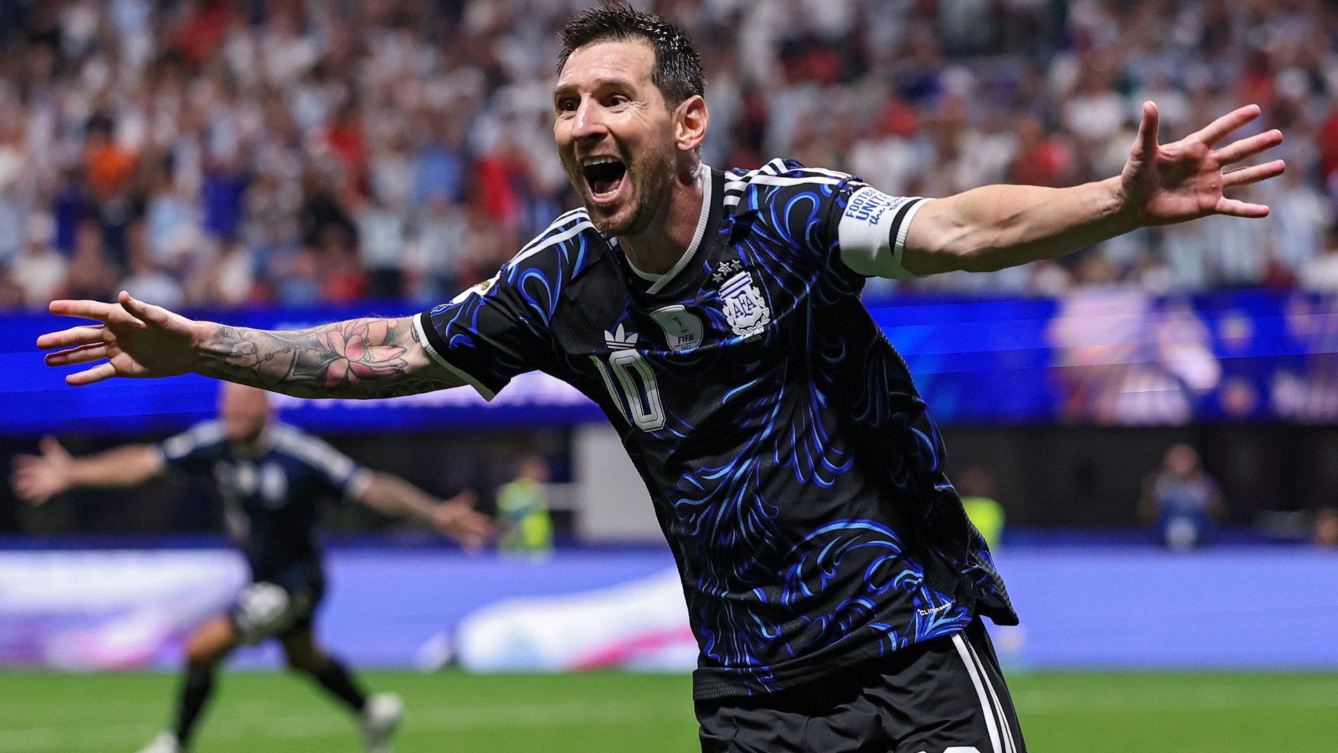 A soccer player wearing a black and blue number 10 jersey celebrates with outstretched arms and an open-mouthed shout in front of a packed stadium crowd.