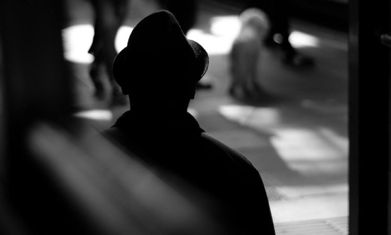 A silhouetted figure wearing a fedora-style hat is seen from behind in a black-and-white photograph, with blurred lights and pedestrians visible in the background.