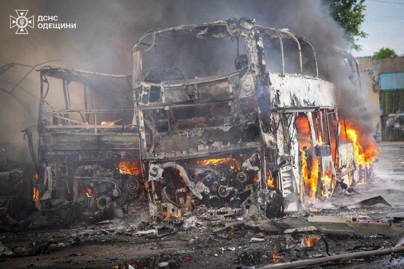 A burned-out bus with shattered windows burns on a debris-strewn street, flames and smoke rising from its wreckage.