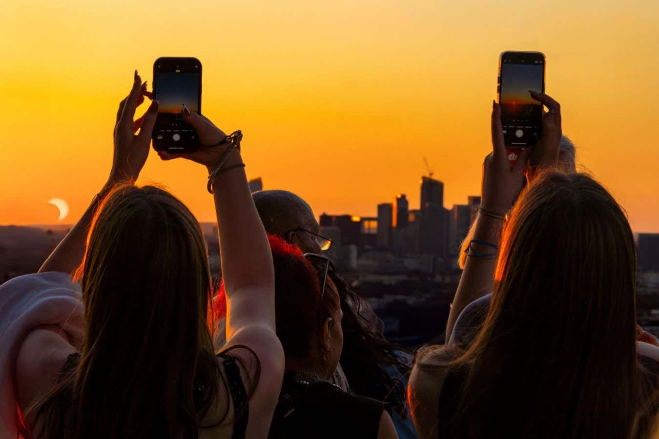 Two people raise smartphones to photograph a partial solar eclipse silhouetted against an orange sunset above a city skyline.