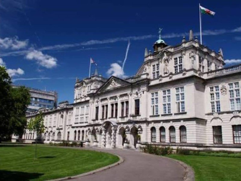 Ornate pale stone building with classical architecture, arched entryway, rooftop statues, and two flags flying under a blue sky, fronted by a manicured lawn.