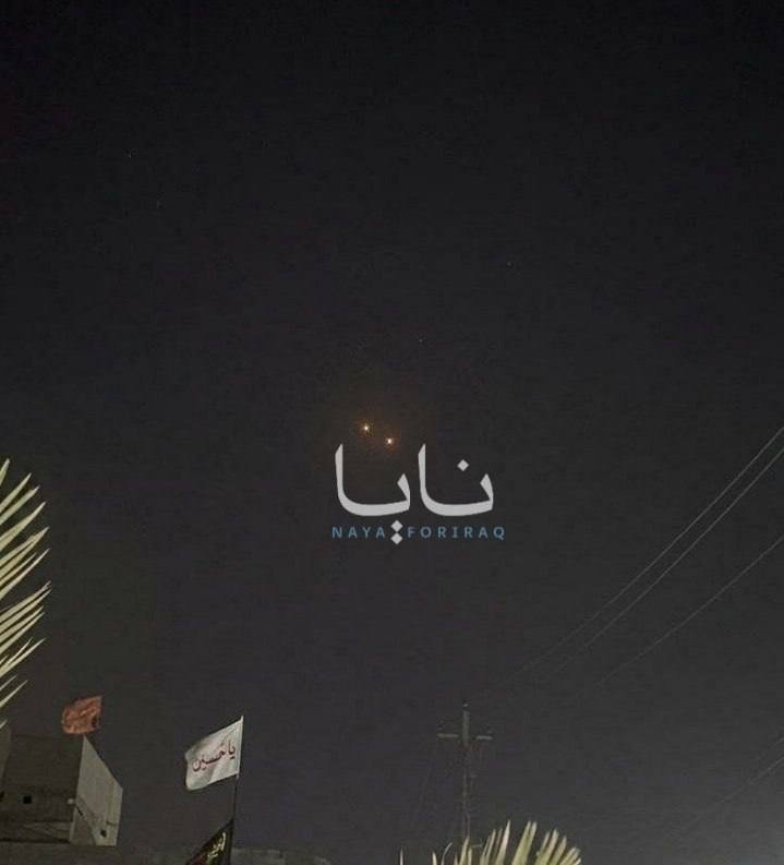 Nighttime scene: two glowing orange lights appear in the dark sky above rooftops with Arabic-script flags and power lines, overlaid with a "NAYA FOR IRAQ" logo.