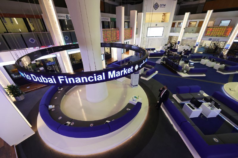 A man in dark attire stands in the expansive, modern interior of the Dubai Financial Market trading floor, featuring circular blue seating arrangements and illuminated digital displays.