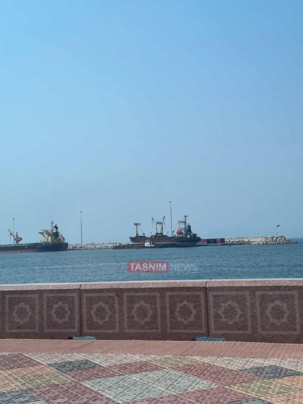 Cargo ships sit docked at a hazy port, viewed from behind a patterned tile promenade with a "TASNIM NEWS" watermark visible on the water.