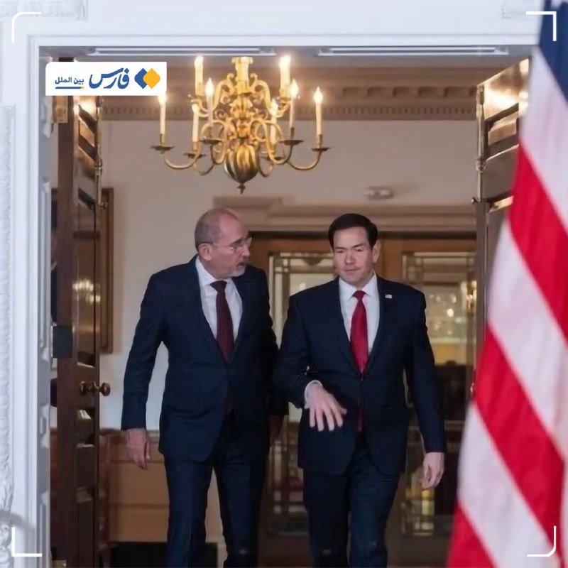 Two men in dark suits and red ties walk side-by-side through a doorway beneath a chandelier, with an American flag partially visible on the right.