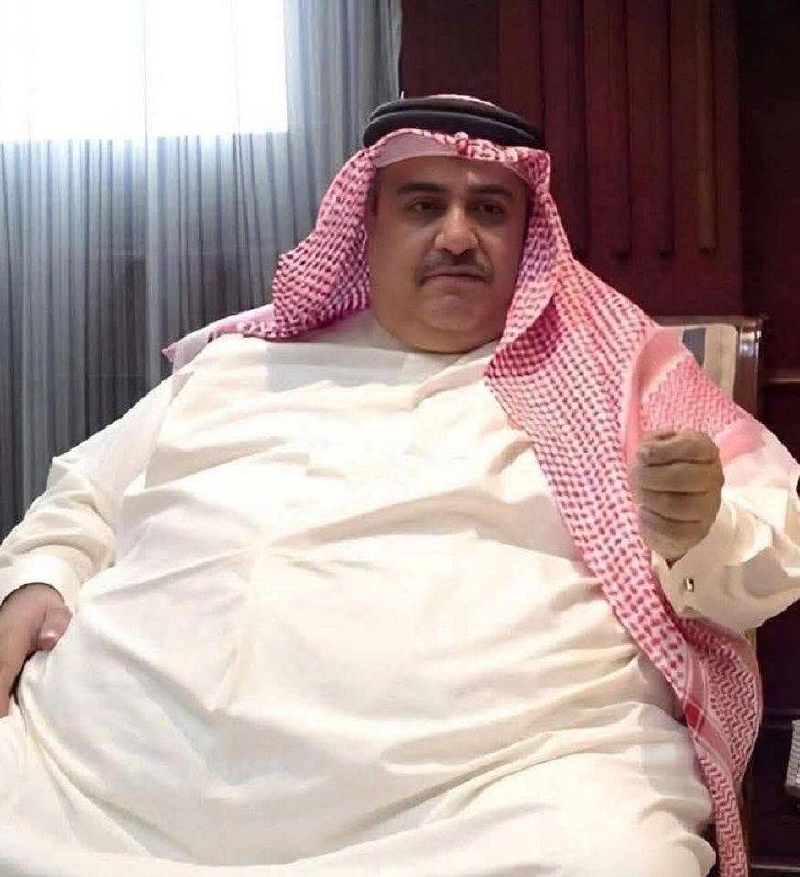 A man wearing a traditional white thobe and a red-and-white checkered keffiyeh with a black agal sits indoors, gesturing with his right hand.