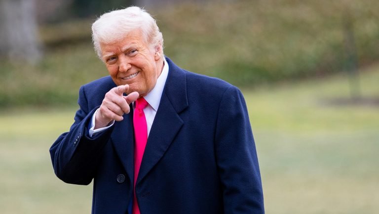 A man with blond hair wearing a navy coat and bright pink tie smiles while pointing his finger toward the camera, standing outdoors on a grassy lawn.