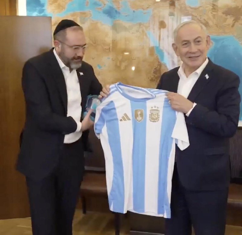 Two men in dark suits hold up a blue-and-white striped Argentina national football team jersey in front of a wall map.