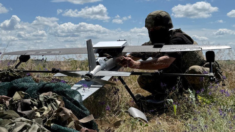 A masked person in military gear kneels in a grassy field, assembling a small fixed-wing drone under a partly cloudy sky.
