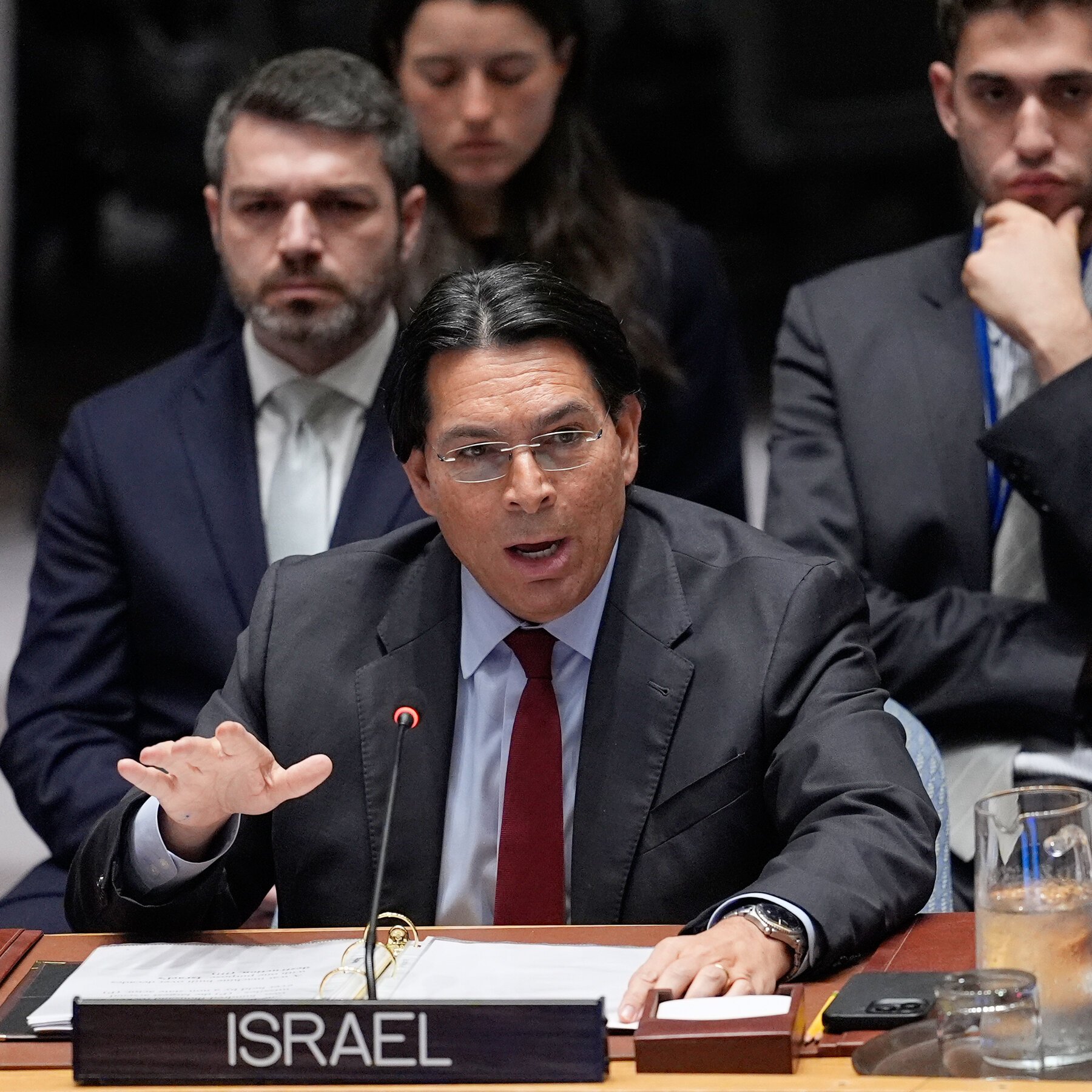 A man in a suit and red tie speaks into a microphone behind an "ISRAEL" placard, with three men seated behind him.