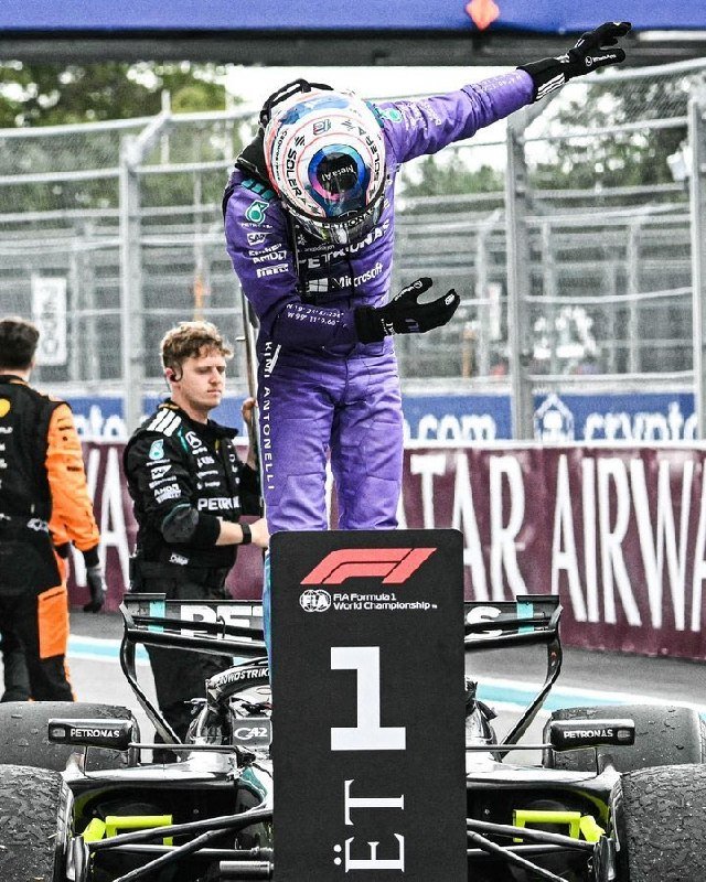 A driver in a purple racing suit stands atop a Formula 1 car marked "1st," waving with both arms raised.