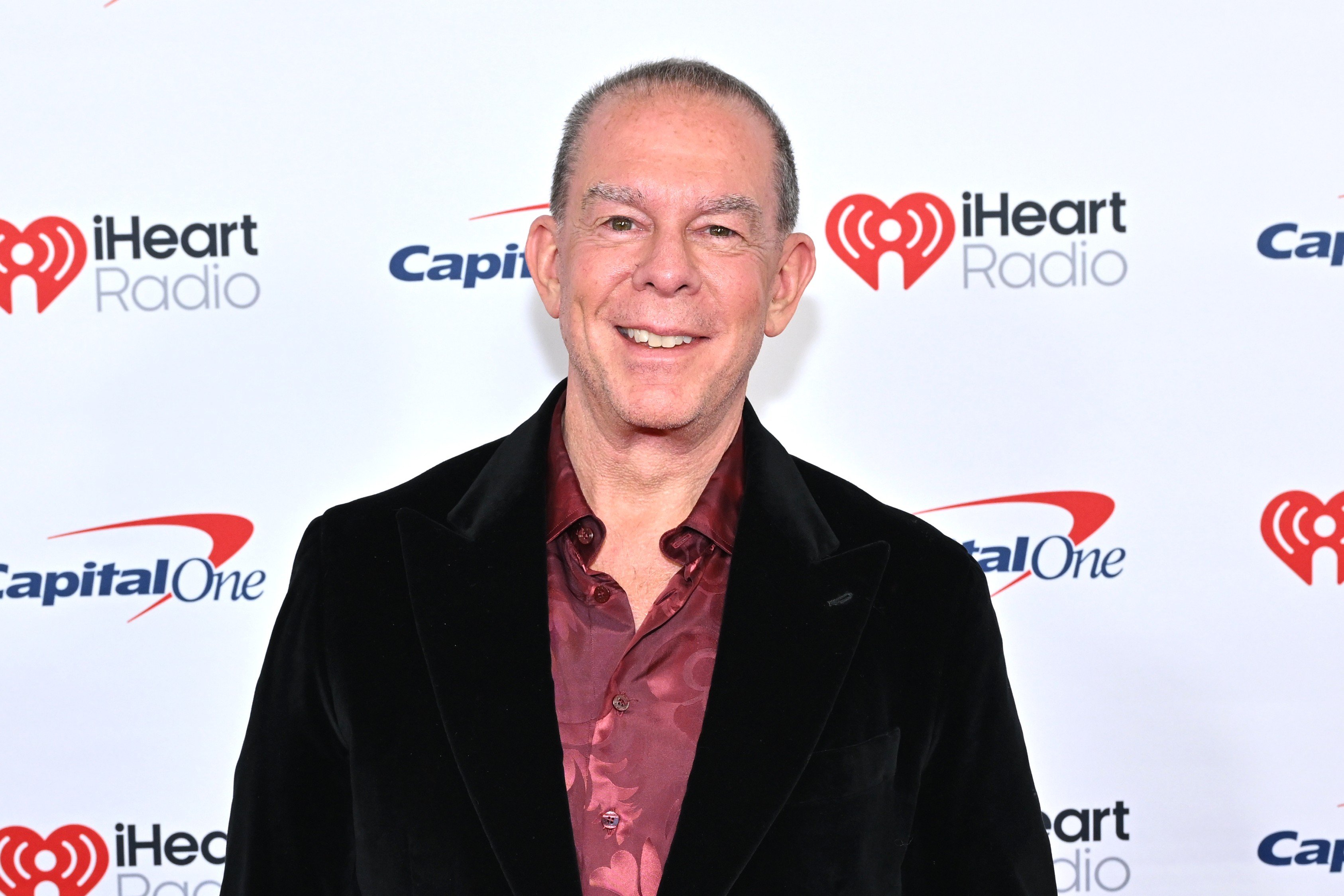 A smiling older man in a black velvet jacket and burgundy patterned shirt poses on a red carpet in front of an iHeartRadio and Capital One backdrop.